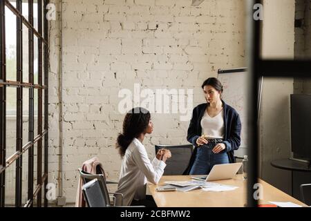 Due attraenti donne multietniche imprenditori che lavorano su un nuovo progetto in un ufficio moderno, seduti alla scrivania Foto Stock