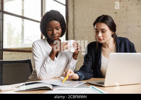 Due attraenti donne multietniche imprenditori che lavorano su un nuovo progetto in un ufficio moderno, seduti alla scrivania Foto Stock