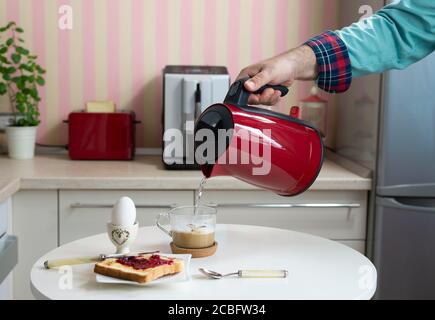 La colazione viene servita su un tavolo da cucina - uova sode e. pane e marmellata con una mano porendo acqua calda da un bollitore in una tazza di caffè Foto Stock