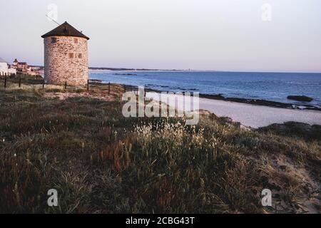 Mulino a vento in pietra sulla spiaggia di Puglia, Portogallo. Vista dell'oceano atlantico da una duna erbosa. Foto Stock