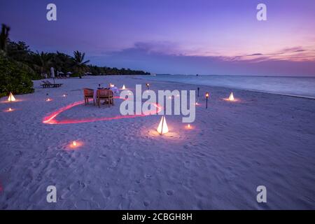 Una giovane coppia condivide una cena romantica con il cuore delle candele sulla spiaggia di sabbia del mare. Cena romantica al tramonto con candele cuore sulla spiaggia di sabbia del mare cielo Foto Stock