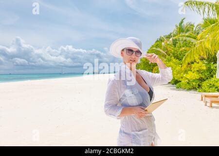 Donna che usa un computer tablet in vacanza in un resort di lusso, occhiali da sole e abito bianco con cappello. Relax ricreazione e chaise lounge, viaggio di piacere Foto Stock
