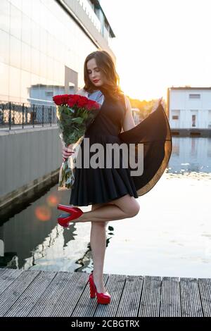 Vista completa della giovane donna con i capelli marroni in abito nero poco che posa e tenendo un bouquet di rose rosse sullo sfondo del restau Foto Stock