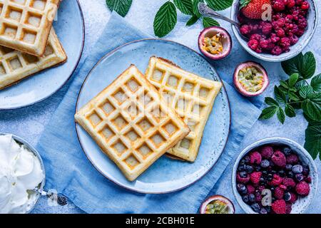 Waffle belgi con lamponi, mirtilli e frutto della passione. Sul tavolo c'è una ciotola di panna montata. Il cibo è visto dall'alto, piatto posato Foto Stock