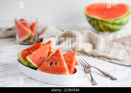 Una ciotola di fette di anguria con forchette sul tavolo. Sullo sfondo c'è un cocomero a metà, un bicchiere con qualche pezzo di melone e una linea Foto Stock