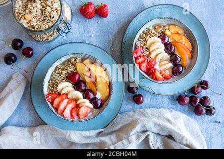 Una sana colazione a base di yogurt con frutta fresca, muesli e quinoa soffiato. Le ciotole grigie blu sono riempite di fragole, pesche, banane e ch Foto Stock