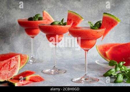 Tre bevande fresche e surgelate di margarita con fragole e cocomero. Le bevande sono guarnite con foglie di basilico e anguria. Sfondo grigio Foto Stock
