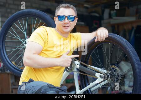 un ragazzo con gli occhiali e una t-shirt gialla si siede dentro parte anteriore di una bicicletta sullo sfondo di un'officina garage Foto Stock