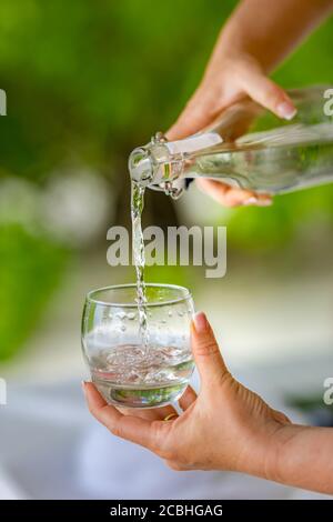 Versare acqua. Primo piano versare acqua fresca purificata dalla bottiglia sul tavolo su uno sfondo naturale sfocato Foto Stock