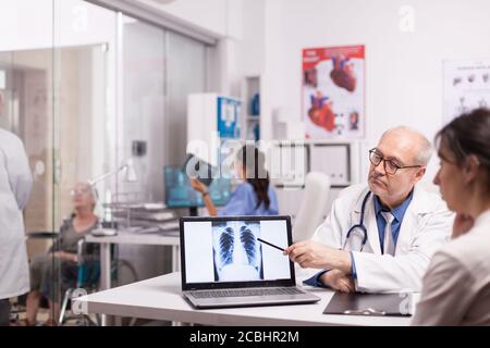 Medico senior che indica la radiografia dei polmoni sani su un computer portatile in ospedale durante la visita medica del paziente. Giovane medico che spiega la diagnosi per disabilitare la donna matura in sedia a rotelle. Foto Stock