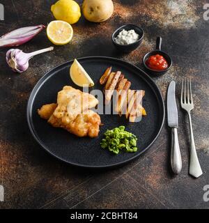 pesce e patatine con baccalà e patatine fritte alla birra e un lato di salsa tartara e piselli funghi su piatto nero sopra il vecchio stile rustico metallo di fondo top Foto Stock