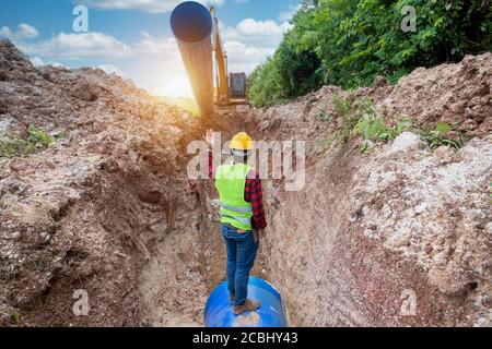 Il tecnico deve indossare l'uniforme di sicurezza esaminando il tubo di drenaggio dello scavo e l'impianto idraulico di grandi dimensioni interrato nel cantiere. Foto Stock