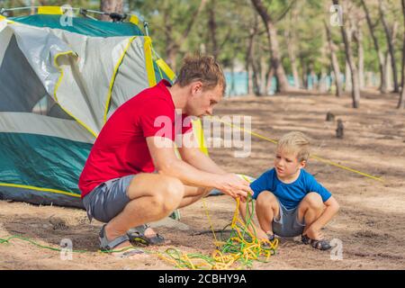 Camping gente stile di vita all'aperto turisti mettendo in su la messa a punto il loro campeggio grigio verde nella foresta estiva vicino al mare di lazur. Carino figlio ragazzo gli aiuta Foto Stock