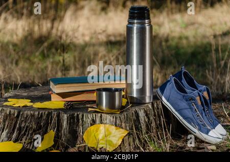 Libri, bottiglia di thermos e una tazza di caffè o tè, sneakers di denim senza lacci e foglie gialle caduti su un ceppo nella foresta o nel parco autunnali. All'aperto Foto Stock