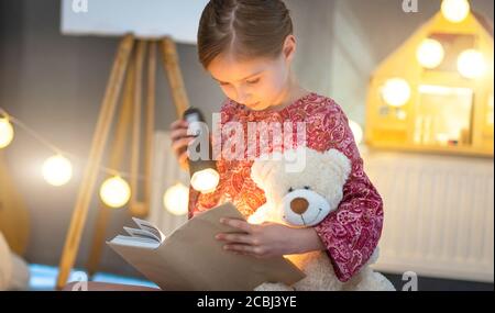 Carino libro di lettura ragazza piccola con torcia in stanza illuminata Foto Stock