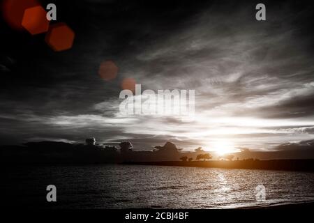 tramonto su cielo nuvoloso alla spiaggia di mare con silhouette ponte di roccia e sfondo di alberi, colore bianco e nero con effetto flare lente rossa Foto Stock