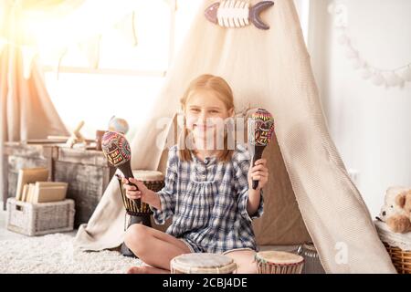 Felice bambina che gioca su maracas di legno in camera decorata con batteria etnica e wigwam Foto Stock