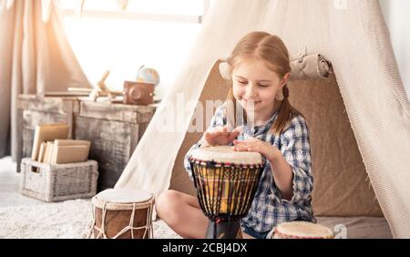 Piccola ragazza batterista che gioca su djembe seduto di fronte tenda etnica a casa Foto Stock