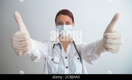 Un medico femminile con una maschera chirurgica e guanti di gomma dà pollici in su. Foto Stock