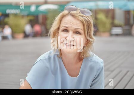 Ritratto di bella donna caucasica invecchiata circa 60 anni con capelli biondi e occhiali da sole. Una donna anziana bianca sta camminando nel parco pubblico. Foto Stock