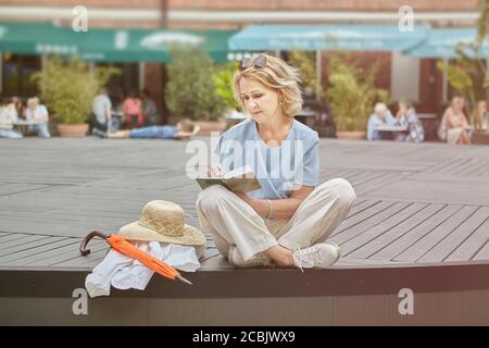 Attiva caucasica anziana circa 60 anni è seduta nel parco pubblico e la lettura di un libro. Donna bianca invecchiata in casual ed elegante abbigliamento passeggiate o Foto Stock