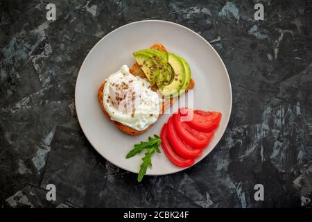 Tostare con uova in camicia, semi di lino, avocado e fette di pomodoro, foglie di rucola su un piatto. Pasto sano per la colazione. Foto Stock