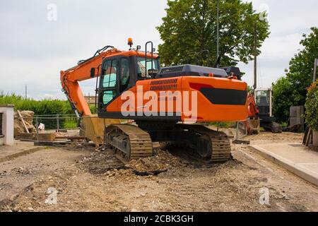 Escavatore cingolato con piattaforma girevole e continuo cingolatura caterpillar su un cantiere di sostituzione fognario a nord Italia occidentale Foto Stock