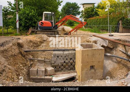 Una trincea per pozzi fognari e un escavatore cingolato compatto con piattaforma rotante e cingolo caterpillar continuo su una fogna Sito di sostituzione in NE Italia Foto Stock