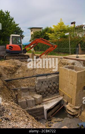 Una trincea per pozzi fognari e un escavatore cingolato compatto con piattaforma rotante e cingolo caterpillar continuo su una fogna Sito di sostituzione in NE Italia Foto Stock