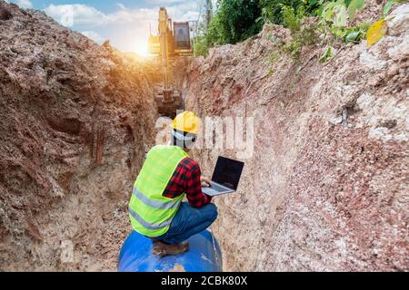 Il tecnico indossa l'uniforme di sicurezza utilizza un computer portatile che esamina il tubo di drenaggio dello scavo e il grande impianto idraulico sotterraneo nel cantiere. Foto Stock