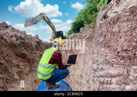 Il tecnico indossa l'uniforme di sicurezza utilizza un computer portatile che esamina il tubo di drenaggio dello scavo e il grande impianto idraulico sotterraneo nel cantiere. Foto Stock