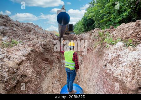 L'uniforme di sicurezza di usura del tecnico tiene un laptop che esamina il tubo di scarico di scavo e l'impianto idraulico grande sotterraneo al luogo di costruzione. Foto Stock