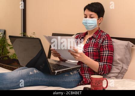 Donna in maschera medica protettiva seduta a letto lavorando a casa su computer portatile durante l'auto-isolamento o la quarantena. Epidemia di coronavirus, epidemia di covid-19 Foto Stock