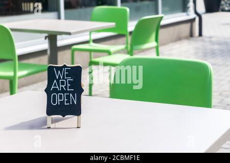 Lavagna con iscrizione aperta sul tavolo nel caffè cittadino. Accesso aperto a mano al bar, all'esterno, per invitare i clienti. Tavoli e sedie del ristorante con Foto Stock