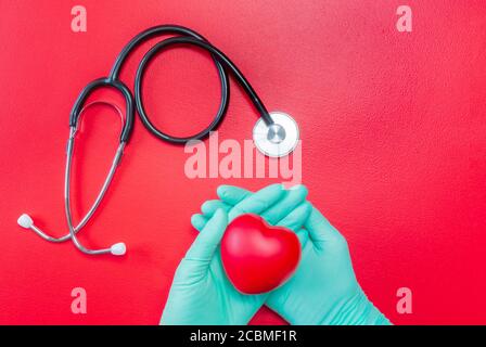 Stetoscopio medico e mani in guanti di gomma che tengono il cuore rosso su sfondo rosso. Posa piatta del cuore nelle mani e nello stetoscopio. Mockup per medico e. Foto Stock