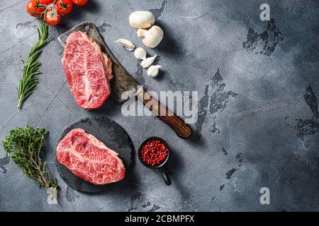 Bistecca di mandrino a lama superiore su ardesia nera e spiedino di macellaio di carne di manzo marmorizzato con erbe pomodori pepe grani su fondo di superficie di pietra grigia Foto Stock