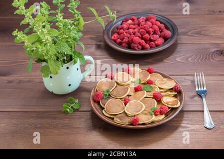 Pancake cereali e lamponi freschi maturi rossi in un piatto marrone. Un mazzo di Melissa in una tazza su un tavolo di legno scuro. Estate Foto Stock