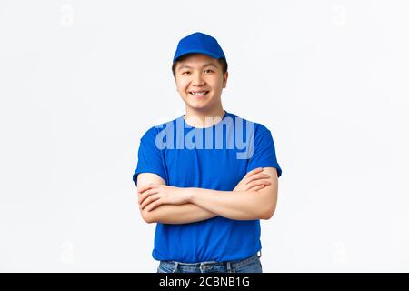 Sorridente ragazzo di consegna asiatico amichevole in blu uniforme braccia trasversali e aspetto fiducioso, in piedi bianco sfondo pronto ad aiutare, portare ordine a. Foto Stock