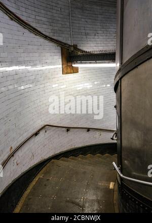 Scala a chiocciola in metallo scuro e sporco illuminata da luce al neon, che conduce al Greenwich Foot Tunnel sotto il fiume Tamigi a Londra Foto Stock