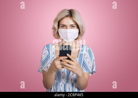 Bella giovane bionda donna con un vestito e una maschera medica sta chattando sul cellulare sorridendo alla fotocamera su un rosa parete dello studio Foto Stock