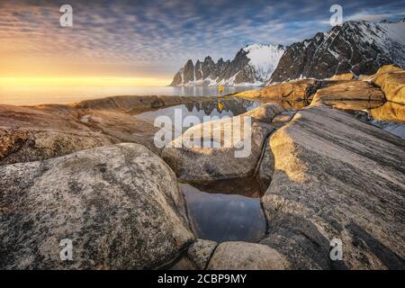 Persona con giacca gialla in piedi sulla spiaggia rocciosa di Toggeneset, riflesso delle cime rocciose innevate Devils denti, Devil's denti Foto Stock