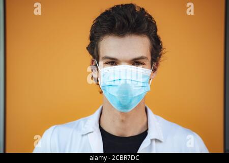 Primo piano ritratto di giovane medico maschile indossando maschera facciale Contro la parete gialla durante l'epidemia di COVID 19 Foto Stock