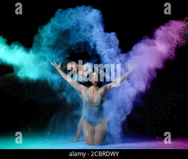 due belle giovani donne caucasiche in tute nere con una figura sportiva ballano in una nuvola di farina viola-blu su uno sfondo nero, esplosi Foto Stock