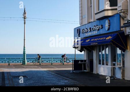 Negozio di fish and chips sul lungomare di Brighton durante la chiusura. Giorno d'estate con il negozio sul lato sinistro, il mare e due ciclisti sullo sfondo. Foto Stock
