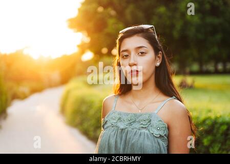 Giovane ragazza latina con lunghi capelli neri e abito blu, con occhiali da  sole sulla testa, al tramonto, con percorso poco focalizzato sullo sfondo,  fissando al ca Foto stock - Alamy