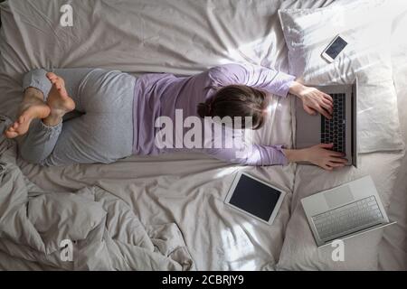 donna sdraiata a letto con tablet digitale e laptop in luce del sole del mattino Foto Stock