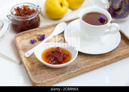 Marmellata di mele cotogne fatta in casa in una ciotola di porcellana con una tazza di tè su un vassoio di legno sullo sfondo è un vaso di vetro con marmellata e mela cotogna. Foto Stock