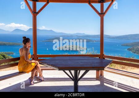 Una donna in abito giallo gode di una vista su Nidri da sotto una rotonda di legno, Lefkada, Isole IONIE, Grecia Foto Stock