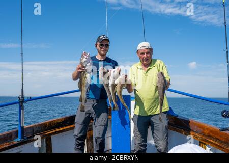 Malmo, Svezia - 6 agosto 2020: Due uomini in un viaggio di pesca mostrano la loro cattura fresca di merluzzo. Oceano blu e cielo sullo sfondo Foto Stock