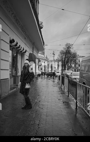 Ragazza in un cappotto di pelliccia. Lviv/Ucraina - 30 gennaio 2020: Una ragazza sta aspettando sul marciapiede bagnato sulla via Petra Doroshenka nel freddo giorno d'inverno Foto Stock